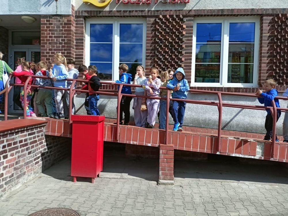 W piątek uczniowie klasy 1a wybrali się na wyjątkową wycieczkę do pobliskiego urzędu pocztowego. Dzieci miały okazję z bliska przyjrzeć się pracy pocztowców, dowiedzieć się, jak wygląda droga listu od nadawcy do adresata, a także zobaczyć, jak działa okienko pocztowe czy stemplowanie przesyłek. Najważniejszym punktem wizyty było jednak coś naprawdę wyjątkowego – uczniowie samodzielnie wysłali listy-niespodzianki z okazji Dnia Mamy. Każda koperta kryła w sobie serce pełne miłości i wdzięczności dla najważniejszych osób w ich życiu.