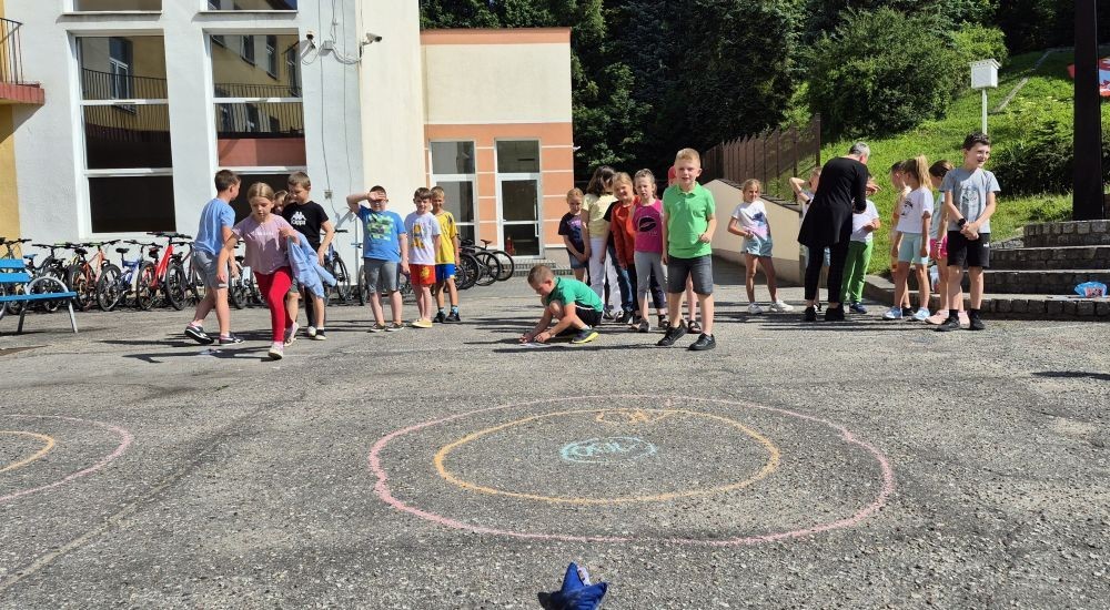 Dziś uczniowie klasy 2b wybrali się na plac przed szkołę, gdzie połączyli przyjemne z pożytecznym. Podczas zabawy w rzuty woreczkiem do celu dzieci nie tylko świetnie się bawiły, ale również ćwiczyły swoją sprawność rachunkową – dodając zdobyte przez grupy punkty.  Radość, ruch i nauka w jednym – właśnie tak lubimy się uczyć.