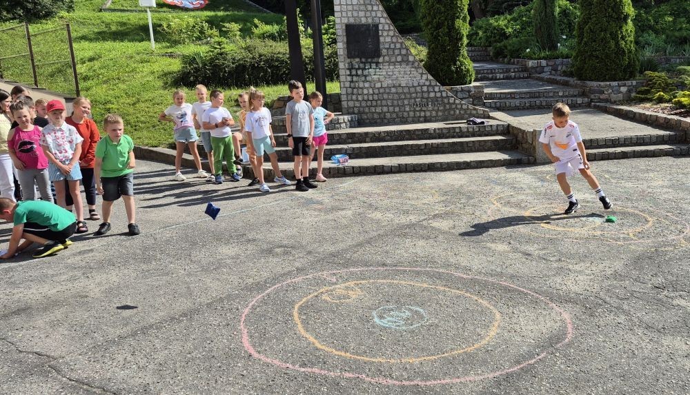 Dziś uczniowie klasy 2b wybrali się na plac przed szkołę, gdzie połączyli przyjemne z pożytecznym. Podczas zabawy w rzuty woreczkiem do celu dzieci nie tylko świetnie się bawiły, ale również ćwiczyły swoją sprawność rachunkową – dodając zdobyte przez grupy punkty.  Radość, ruch i nauka w jednym – właśnie tak lubimy się uczyć.