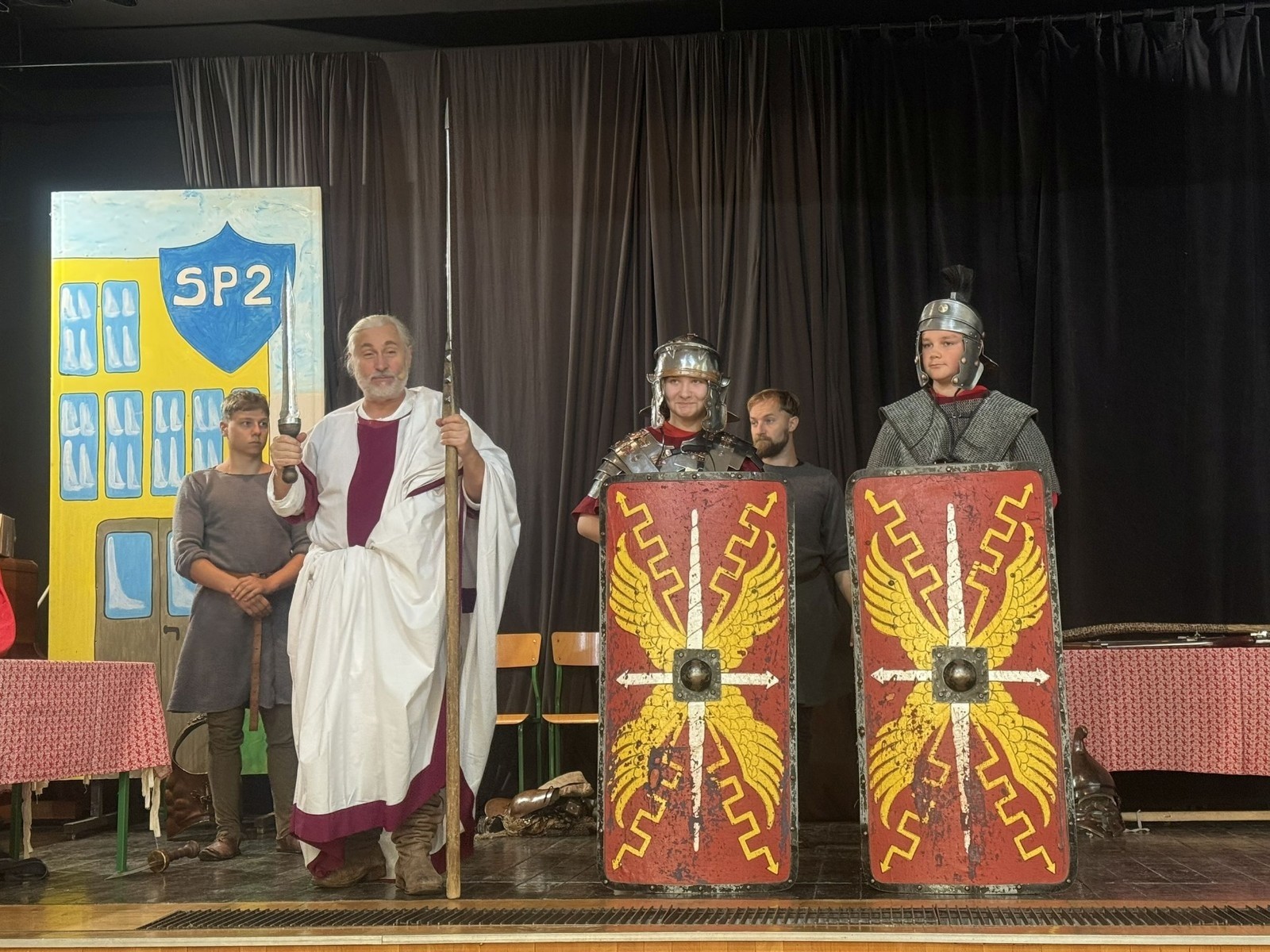 Każdego roku uczniowie naszej szkoły mają niepowtarzalną okazję wziąć udział w wyjątkowym wydarzeniu edukacyjnym - ŻYWEJ LEKCJI HISTORII, prowadzonej przez grupę rekonstrukcyjną Chorągiew Komturstwa Gniewskiego. To spotkanie, które na długo pozostaje w pamięci uczestników, łącząc wiedzę historyczną z emocjami, ruchem i bezpośrednim kontaktem z przeszłością.
W tym roku tematyka spotkań została zrealizowana na dwóch poziomach wiekowych:
Klasy 4-6: Świat starożytnych Greków i Rzymian
Uczniowie klas młodszych przenieśli się do czasów starożytności - epoki potężnych cywilizacji, mitologii, igrzysk olimpijskich i legionów rzymskich. Dzięki strojom z epoki, realistycznym rekwizytom i barwnym opowieściom rekonstruktorów, dzieci mogły poczuć atmosferę antycznego świata. Dowiedzieli się, jak wyglądało życie codzienne Greków i Rzymian, jakie były zwyczaje wojenne, a nawet jak wyglądały ćwiczenia wojskowe legionistów. Zajęcia - prowadzone w formie interaktywnej - dały uczniom możliwość przymierzenia elementów uzbrojenia oraz wzięcia udziału w krótkich pokazach.
Klasy 7-8: Długi wiek XIX
Starsze klasy uczestniczyły w lekcji poświęconej Długiemu wiekowi XIX, czyli okresowi, który ukształtował współczesny świat. Rekonstruktorzy przybliżyli wydarzenia związane z rewolucjami, walką o niepodległość, przemianami społecznymi i narodzinami nowoczesnych idei. Szczególną uwagę poświęcono wydarzeniom ważnym dla Polski - rozbiorom, powstaniom narodowym i postaciom kluczowym dla naszej historii. Uczniowie mogli zobaczyć repliki broni, a także stroje z XIX wieku. Dla wielu była to okazja do poznania historii w sposób bardziej angażujący niż podręcznik - przez opowieści, prezentacje multimedialne i inscenizacje.
Żywa lekcja historii to nie tylko atrakcja, ale przede wszystkim cenna forma nauki. Bezpośredni kontakt z pasjonatami historii oraz aktywne uczestnictwo w przedsięwzięciu sprawia, że uczniowie lepiej zapamiętują materiał, rozbudzają swoją ciekawość i rozwijają szacunek do dziedzictwa kulturowego.